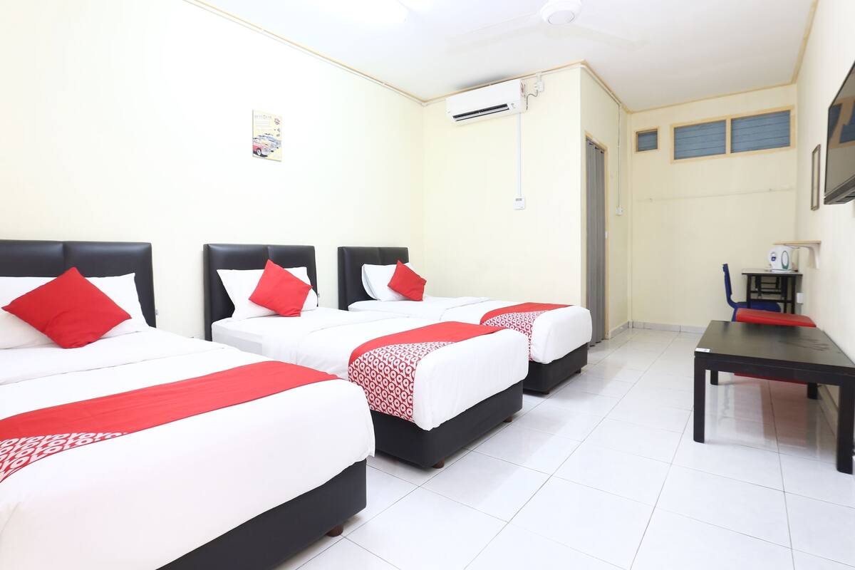 Habitación de hotel con tres camas, cada una con ropa blanca y detalles rojos, un escritorio y un equipo de aire acondicionado en la pared.