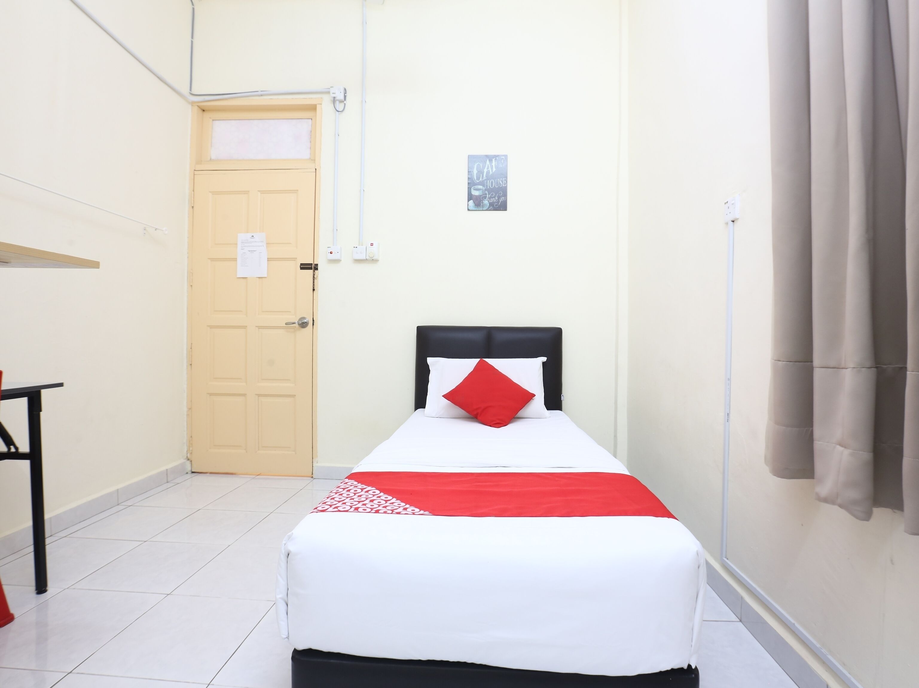 A small, neatly arranged room with a bed, a door, a small table, and a wall-mounted sign.