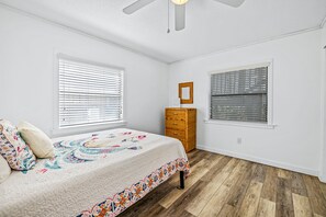 Un dormitorio con cama, dos ventanas con persianas, un tocador de madera y un ventilador de techo.