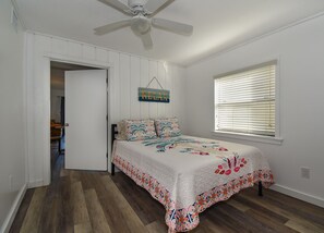 Quarto com cama, ventoinha no teto, janela com persianas e porta que dá para outro cômodo.