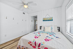A bedroom with a bed, a ceiling fan, a window with blinds, and a wall-mounted sign. - Renovated Sea Dunes 2BR with Ocean View Ground Level and Pool Next Door (New Smyrna Beach)