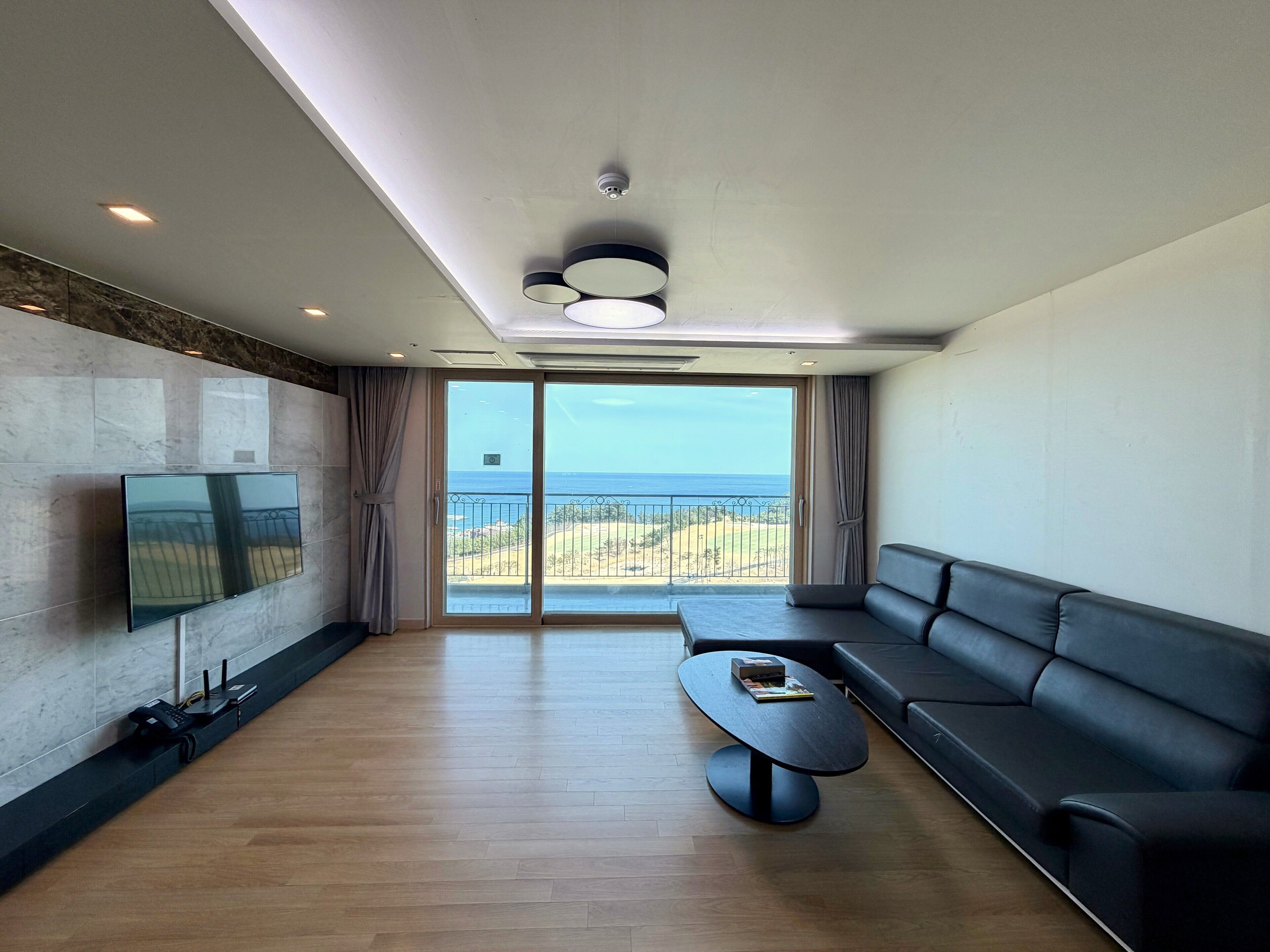 Ein modernes Wohnzimmer mit einem großen Flachbildfernseher an der Wand, einem schwarzen Ledersofa, einem runden Couchtisch und Blick auf den Ozean durch eine Schiebetür.