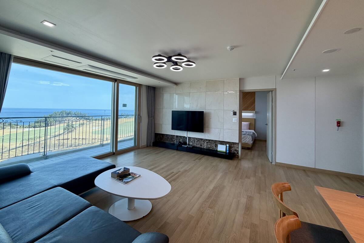 Een moderne woonkamer met een grote televisie, een salontafel en een bank. Er is een balkon met uitzicht op de oceaan.