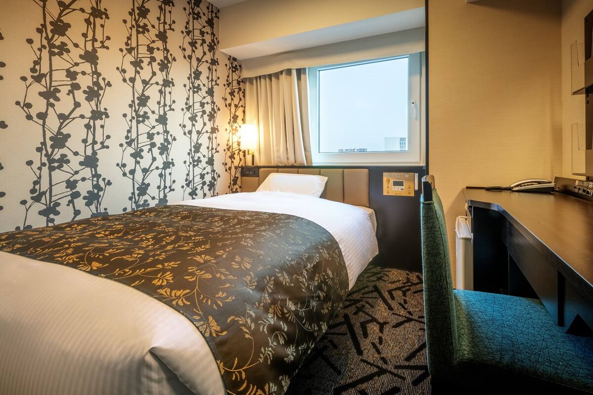 Une chambre d’hôtel comprenant un lit, un bureau, une chaise, une fenêtre et un mur orné d’un motif floral.