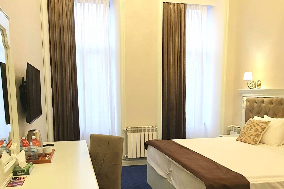 Ein Hotelzimmer mit Bett, Schreibtisch, Stuhl, Fernseher, Heizkörper und einem Fenster mit Vorhängen.