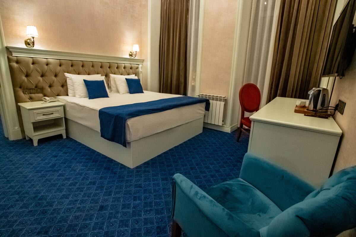 Ein Hotelzimmer mit einem großen Bett, einem Nachttisch, einem roten Sessel, einem Schreibtisch mit Wasserkocher und einem blauen Ohrensessel.
