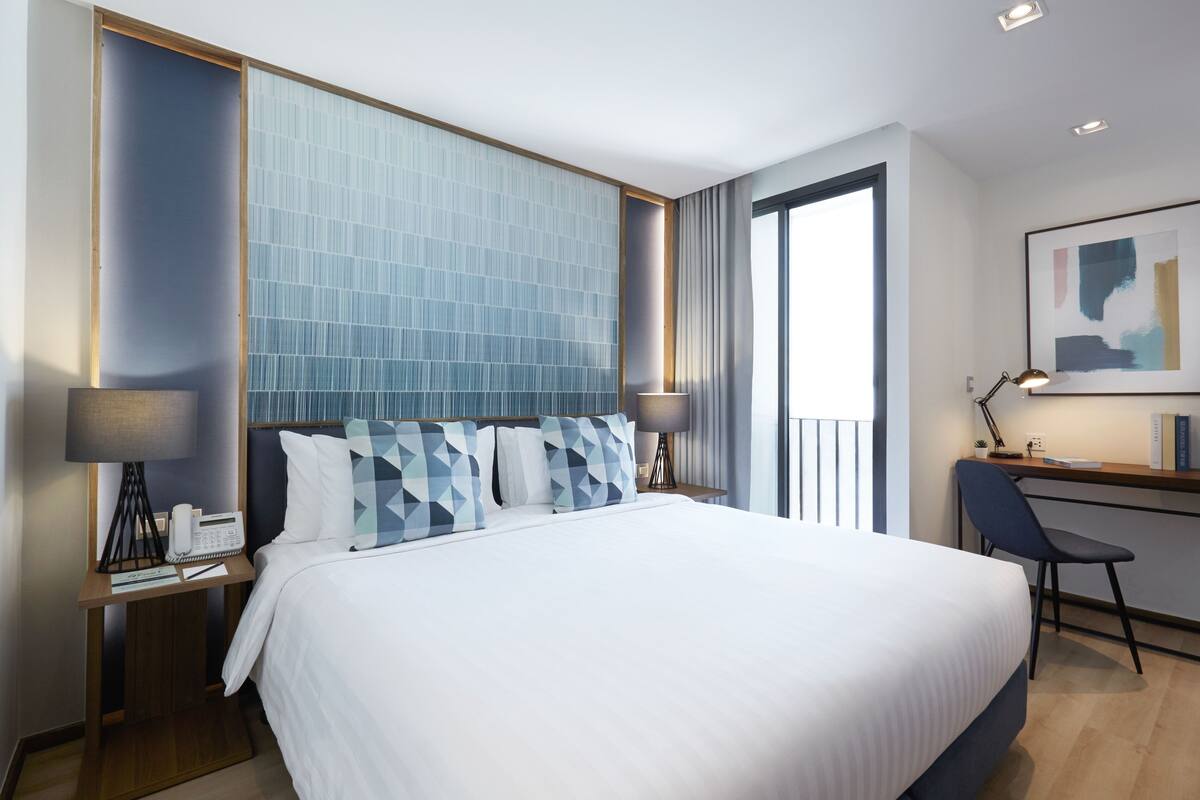 A modern hotel room with a large bed, a desk with a chair, a telephone, and a lamp.