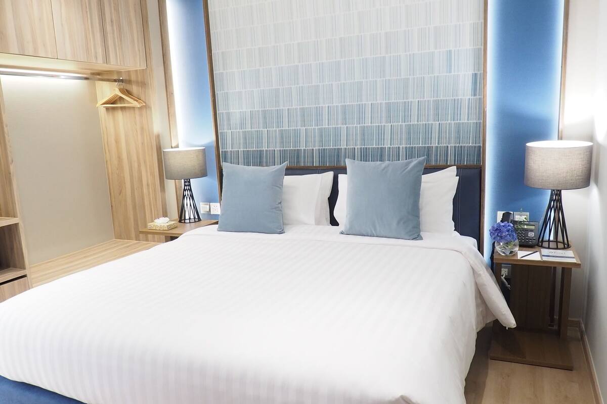 A modern hotel room with a large bed, bedside tables, a wardrobe, and a large window with sheer curtains.