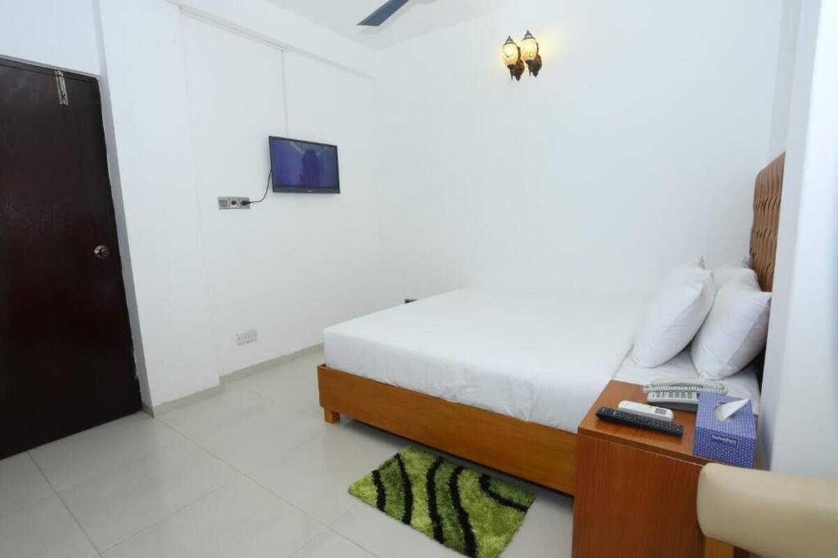 A bedroom with a bed, a TV mounted on the wall, a bedside table with a remote control, and a green rug.