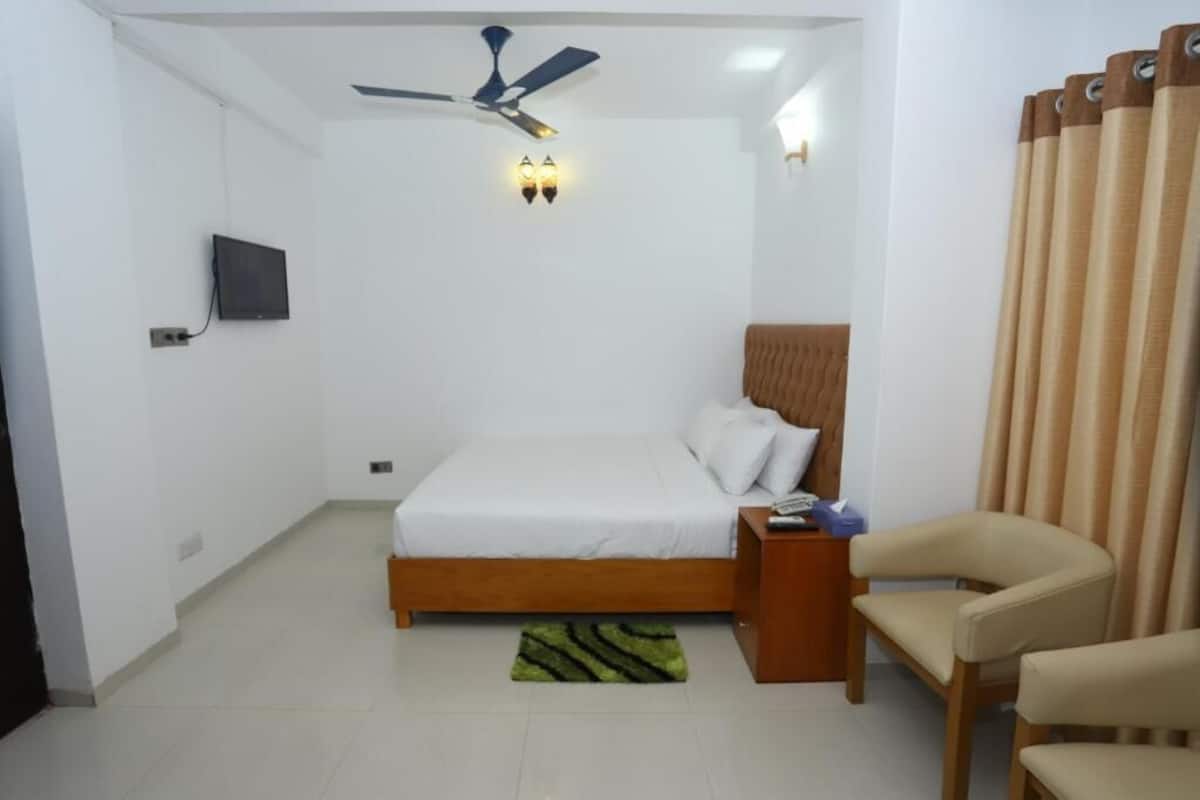 A bedroom with a bed, a ceiling fan, a TV mounted on the wall, a wooden nightstand, a chair, and a window with curtains.