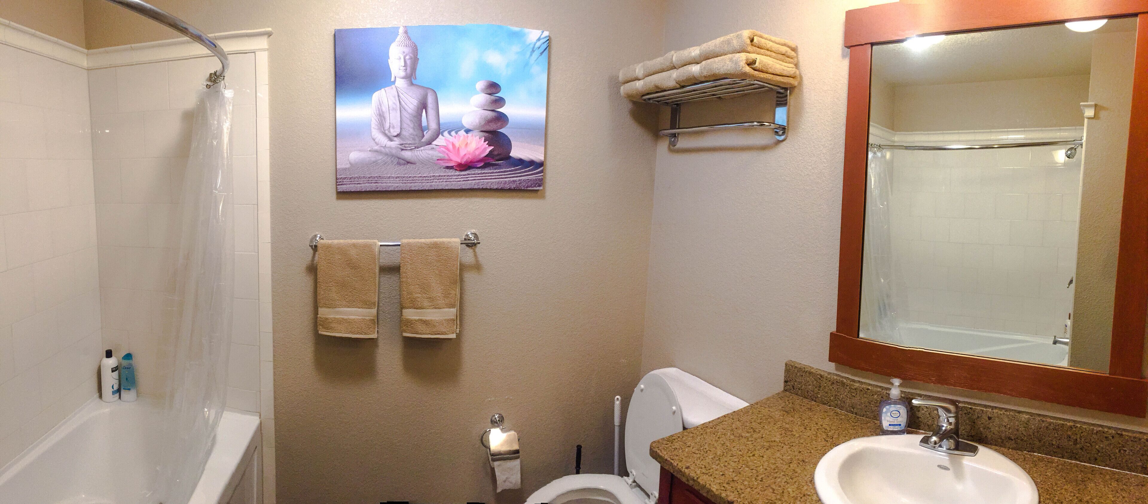Un baño con bañera, inodoro, lavamanos y un espejo grande. Hay un perchero con dos toallas y una imagen de Buda en la pared.