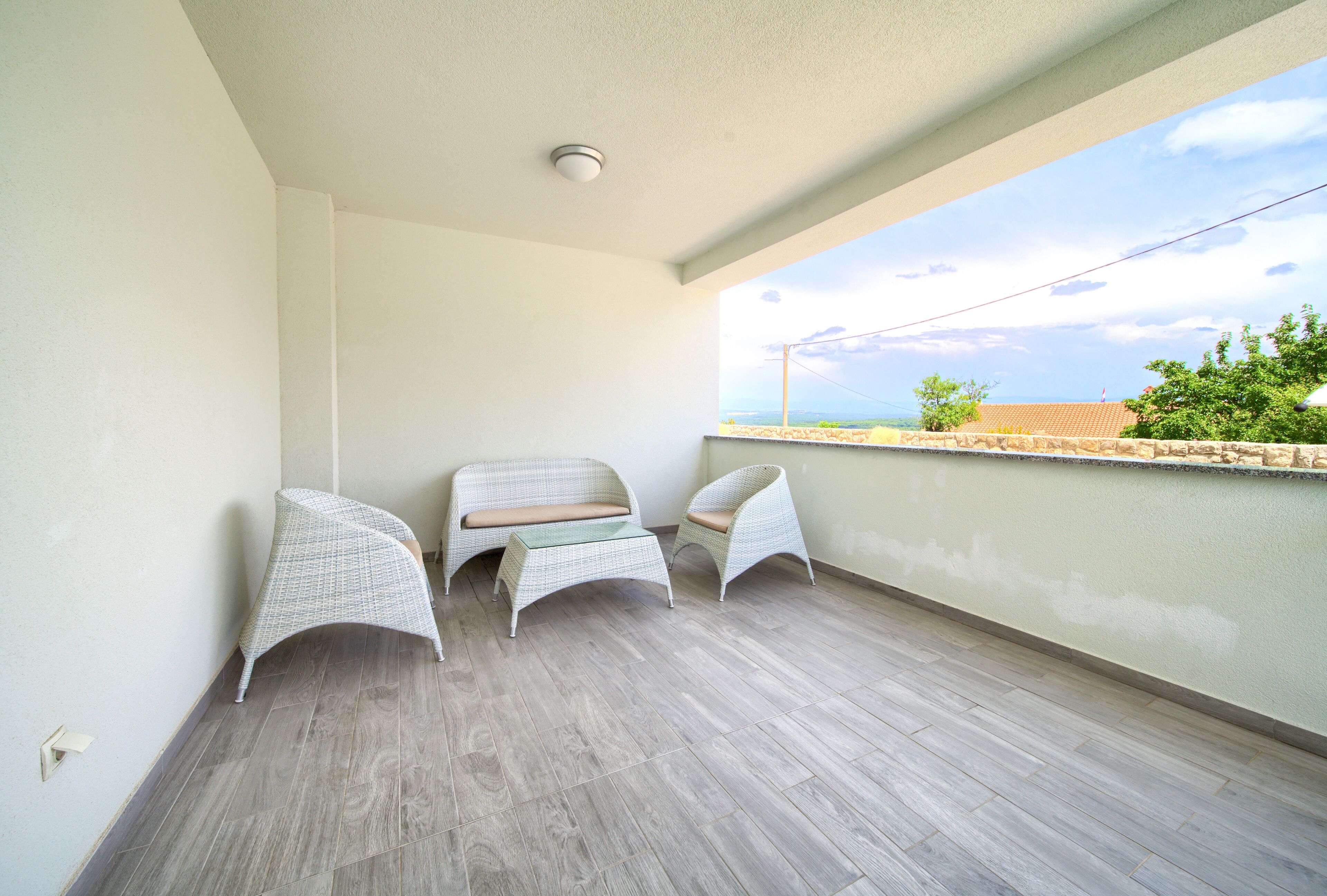 A balcony with two wicker chairs and a small table, a light-colored wall, and a view of the sea and buildings.