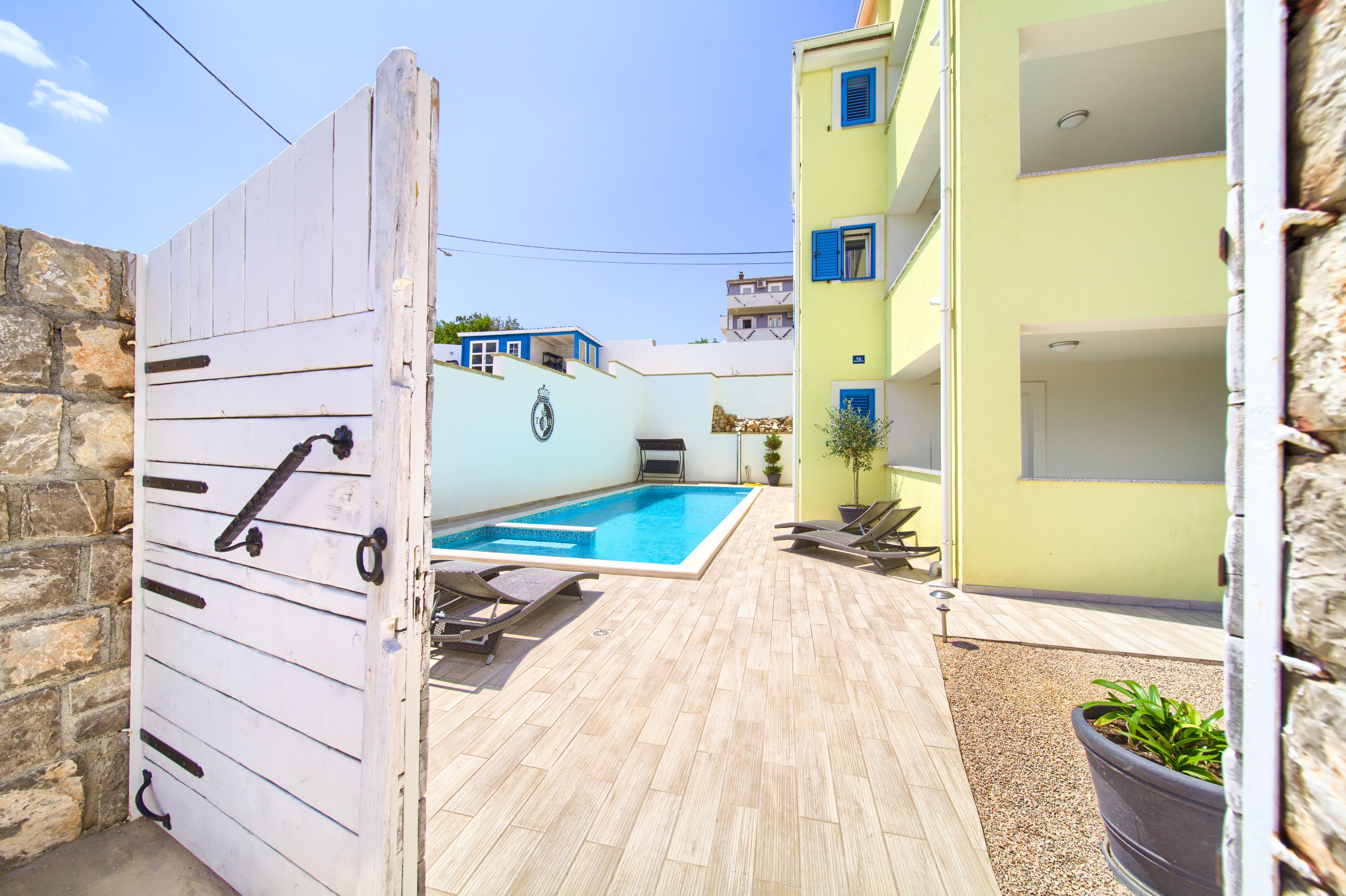 Un espace piscine avec une terrasse en bois, un portail blanc et un bâtiment aux volets bleus.