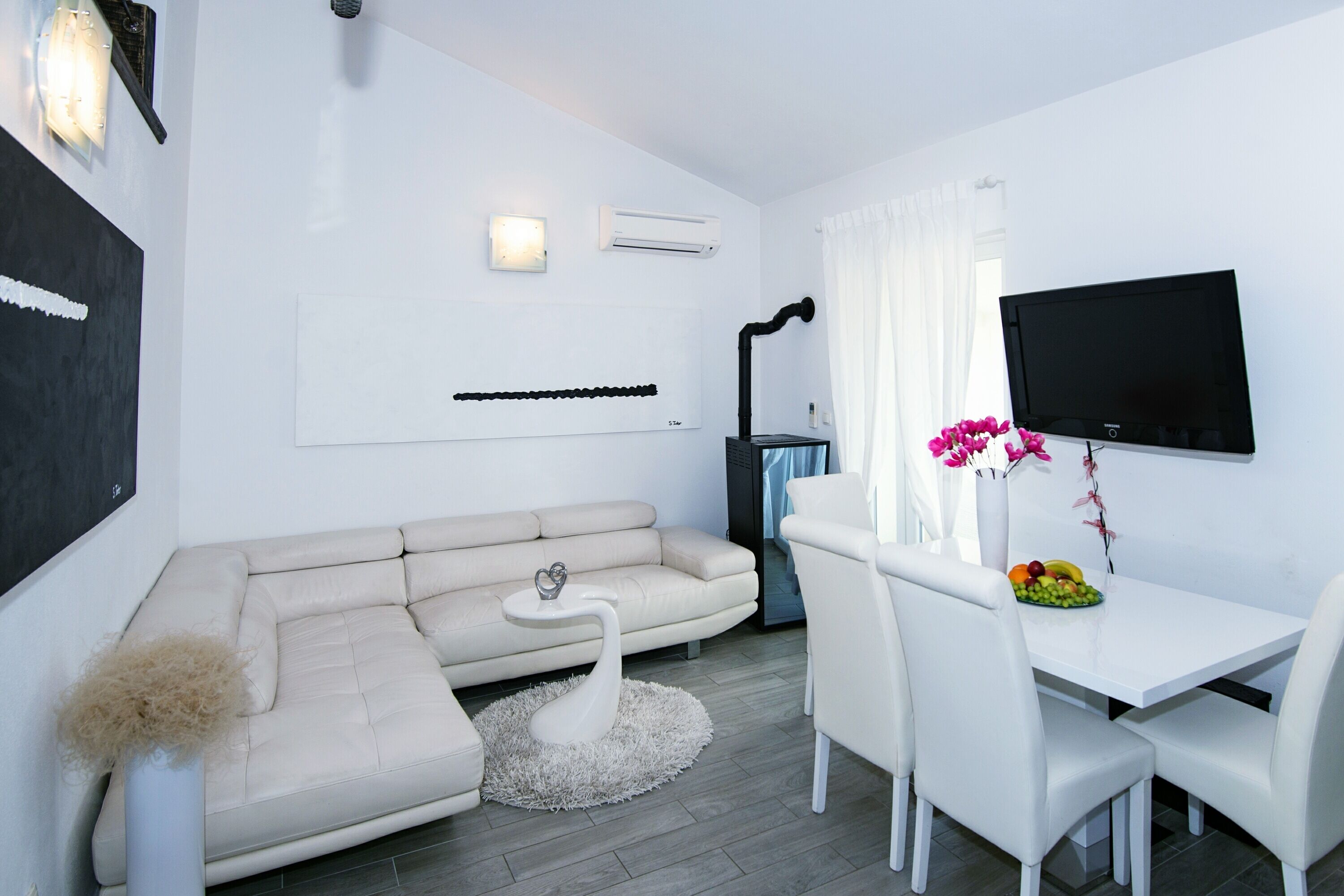 A modern living room with a sofa, a small round table, a white dining table with chairs, a flat-screen TV mounted on the wall, and an air conditioning unit.