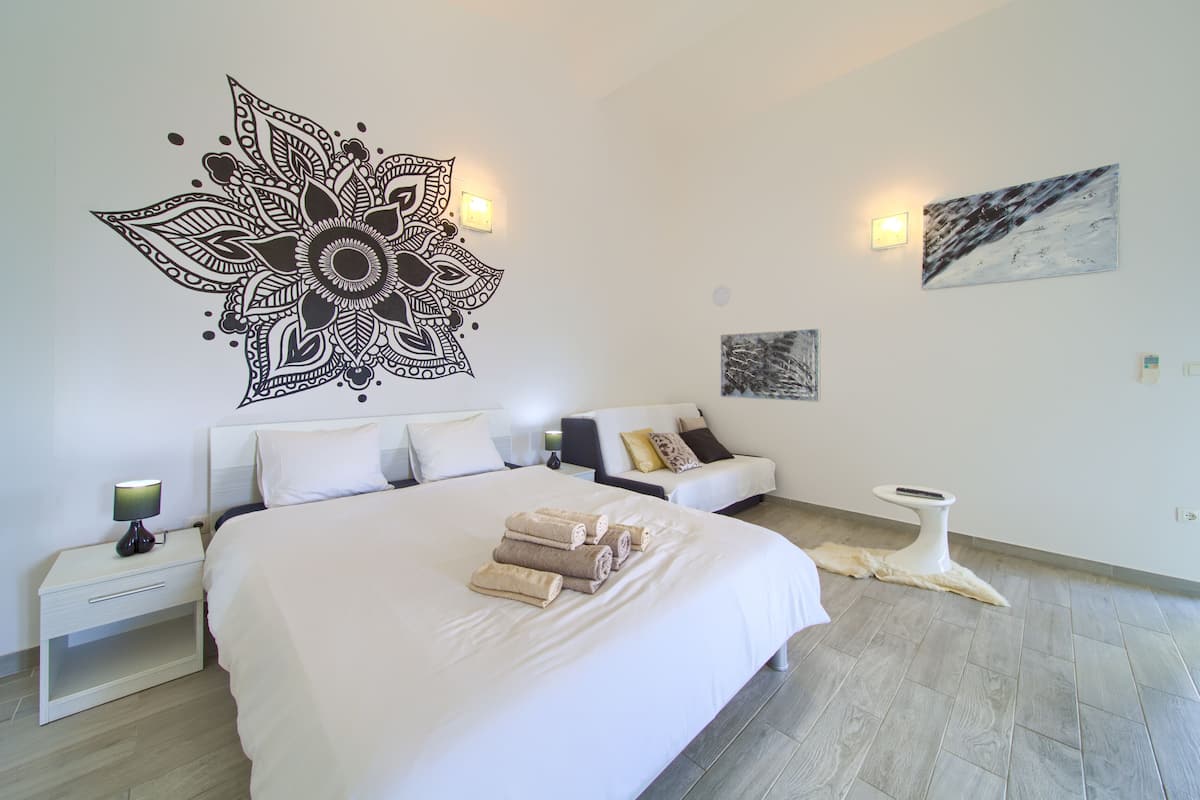 A modern bedroom with a large bed, a nightstand with a lamp, a sofa with cushions, and a wall-mounted artwork.