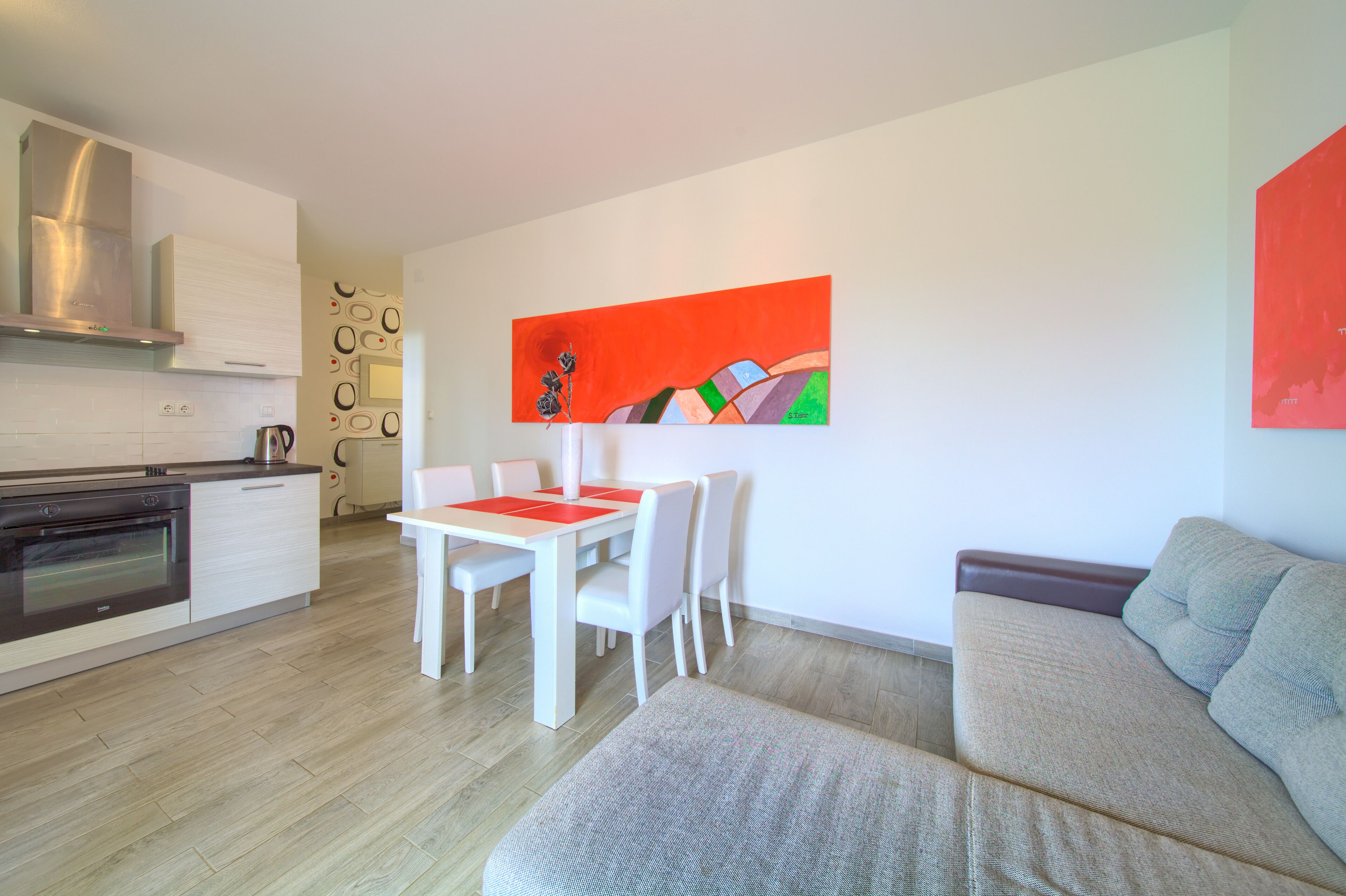 A modern kitchen with white cabinetry and a stainless steel range hood, a dining area with a white table and chairs, and a gray sofa in a living area.