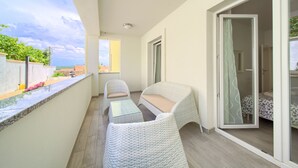 A balcony with wicker furniture, a glass table, and a view of the sky and greenery. - Apartments Mali Nono (Krk)