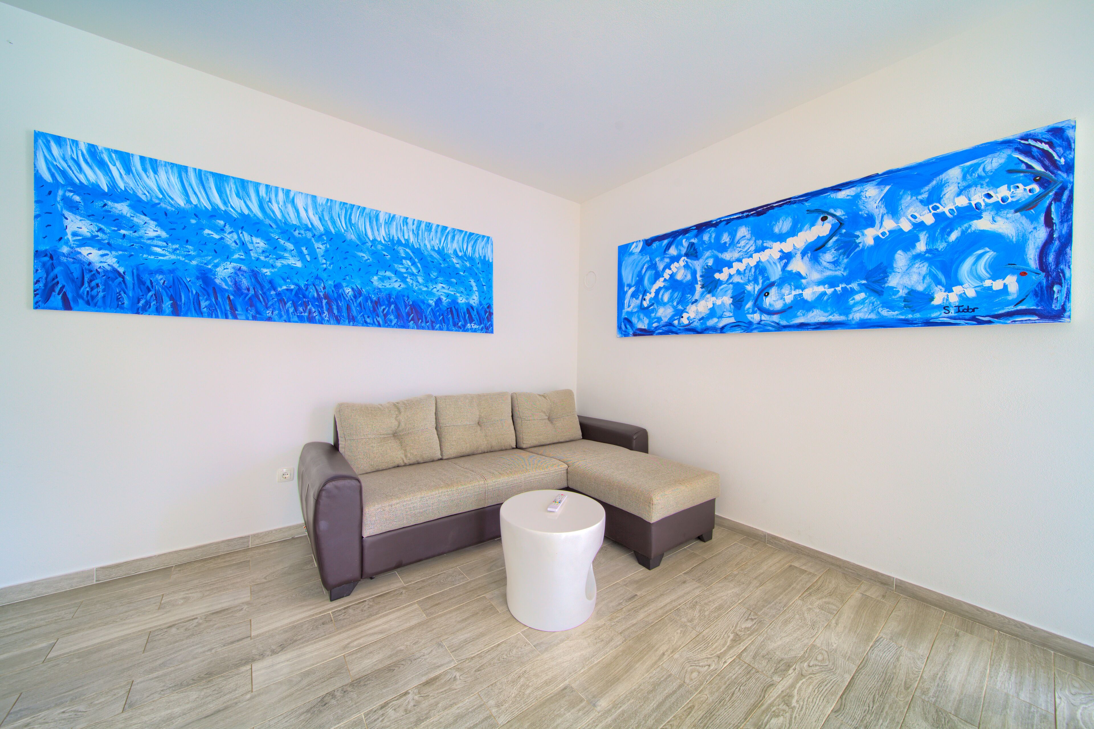 A room with a sectional sofa, a small white table, and two large abstract blue paintings on the walls.