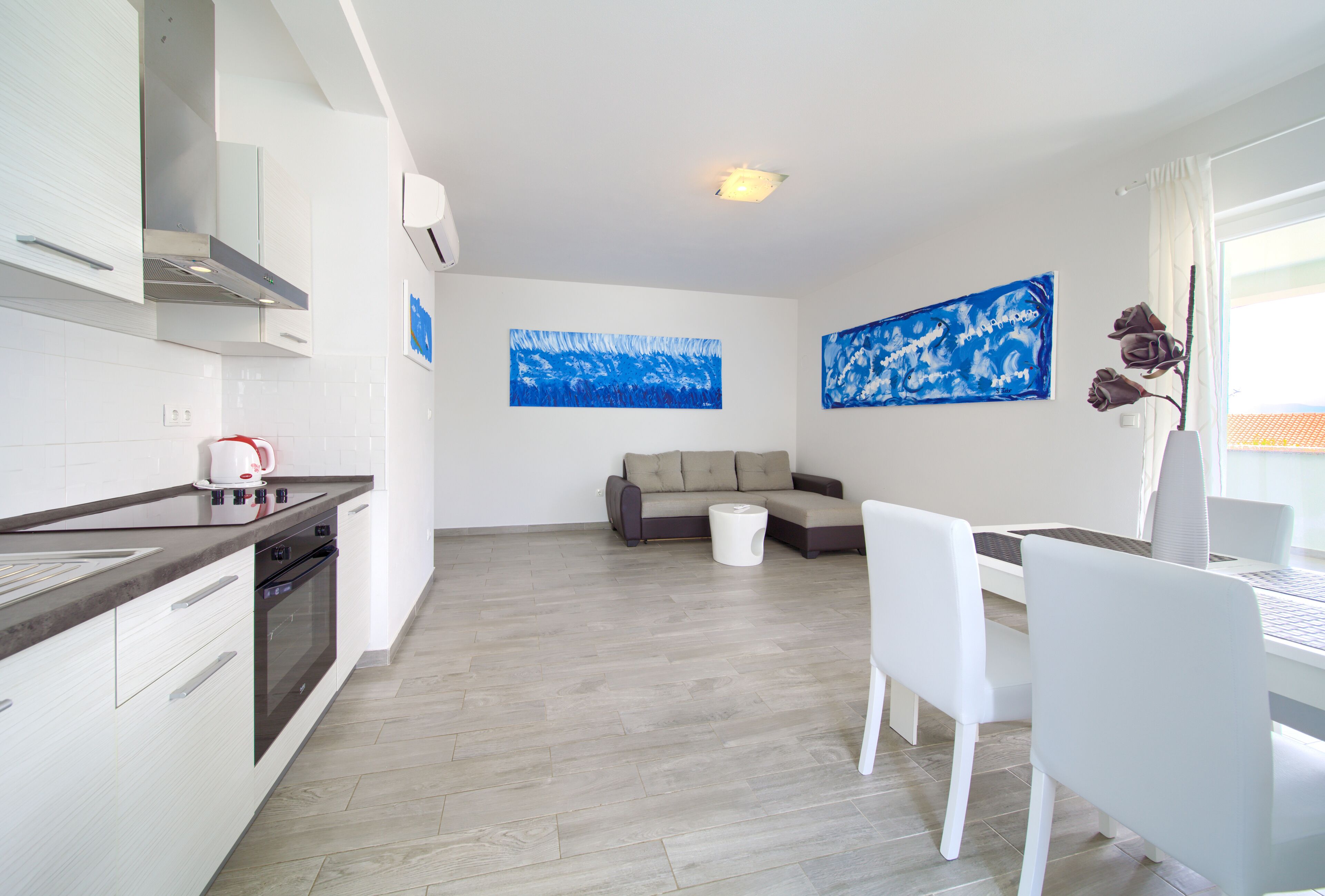 A modern kitchen with white cabinetry, a built-in oven, and a dining area with a white table and chairs. There are two abstract blue paintings on the walls.