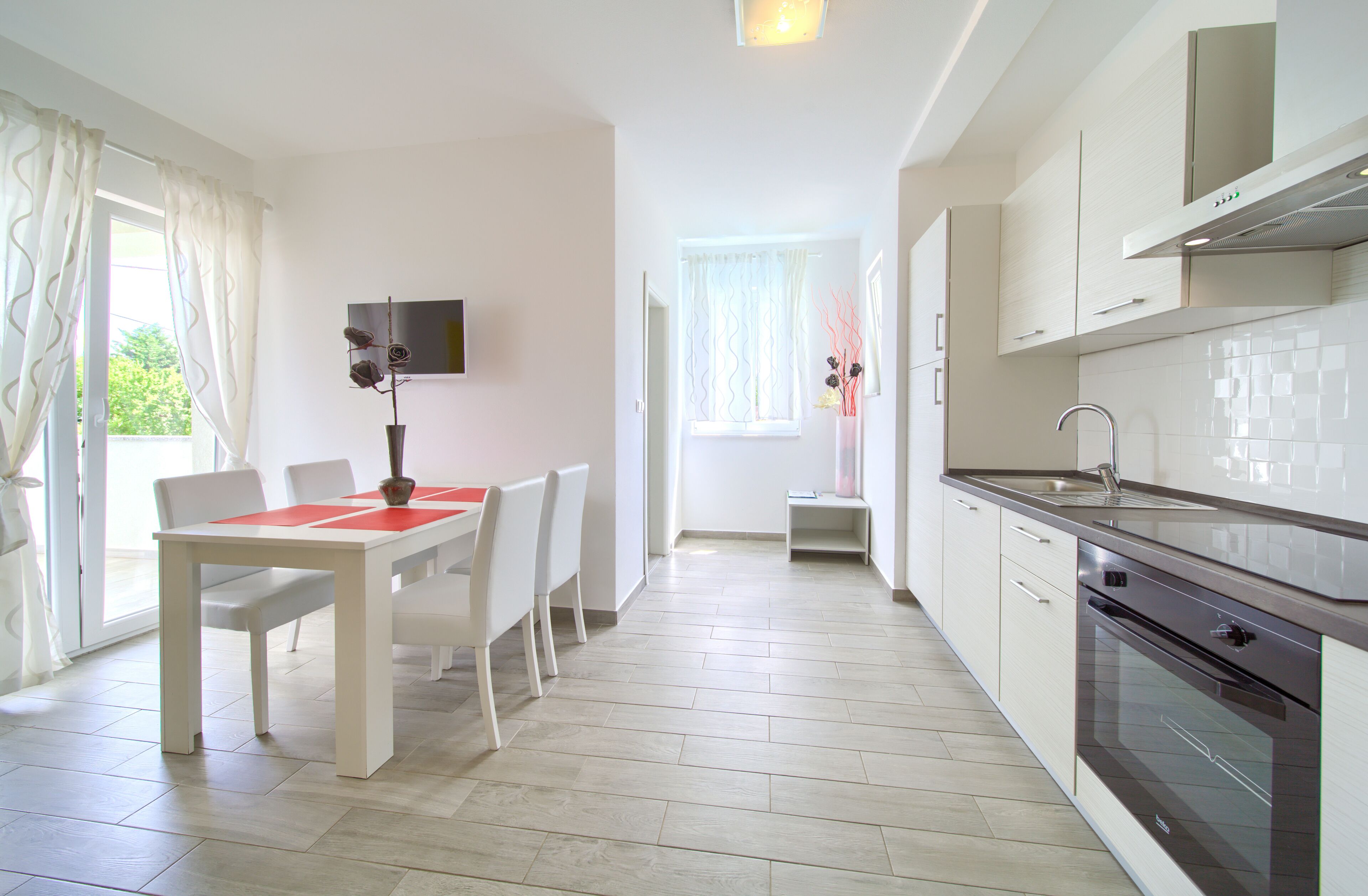 A modern kitchen with white cabinets, a stainless steel sink, and a built-in oven. There is a dining area with a white table and chairs, and a vase with flowers on the windowsill.