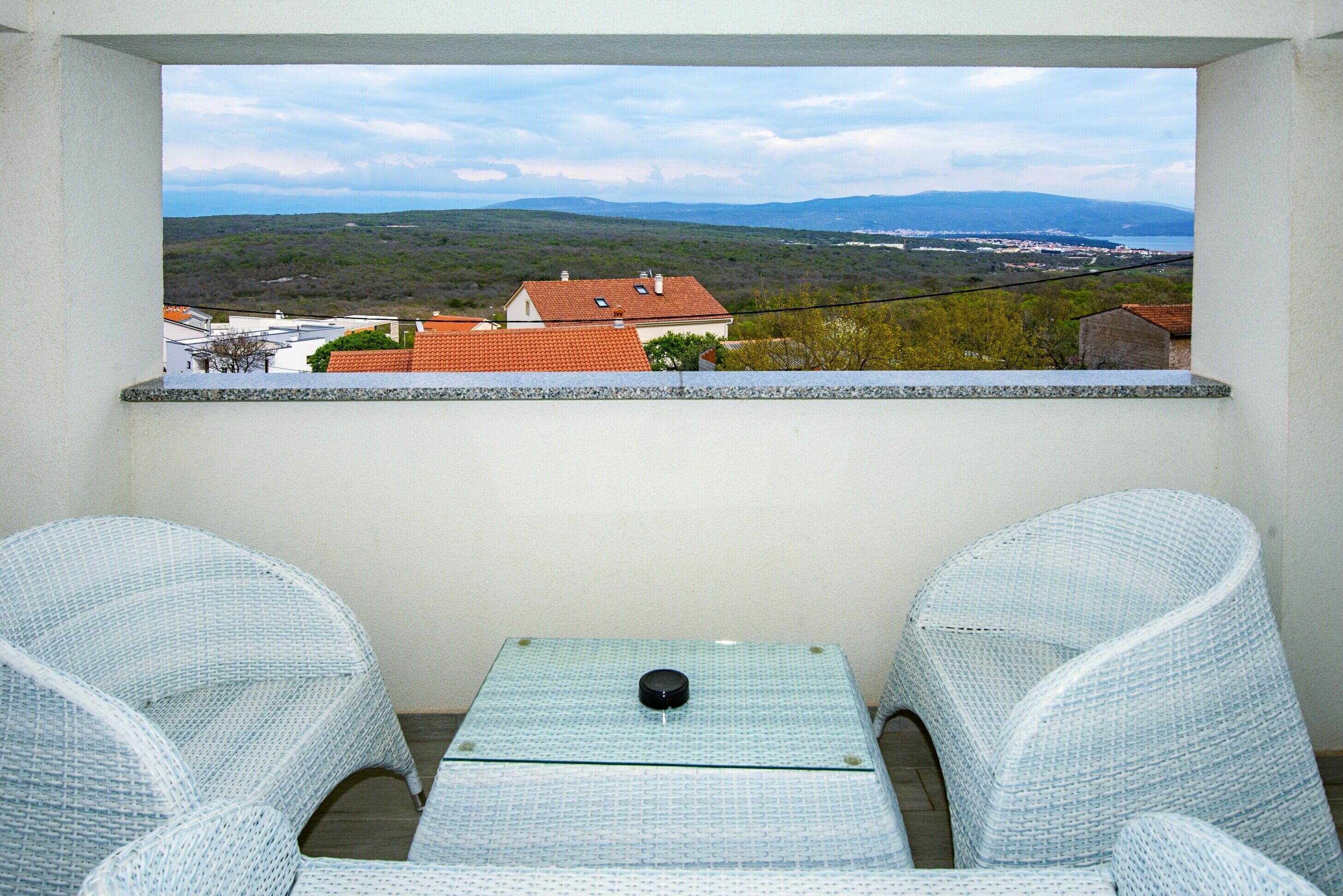 Un balcon agrémenté de fauteuils en osier et d’une petite table, offrant une vue panoramique sur un paysage pittoresque, avec des maisons et des montagnes.