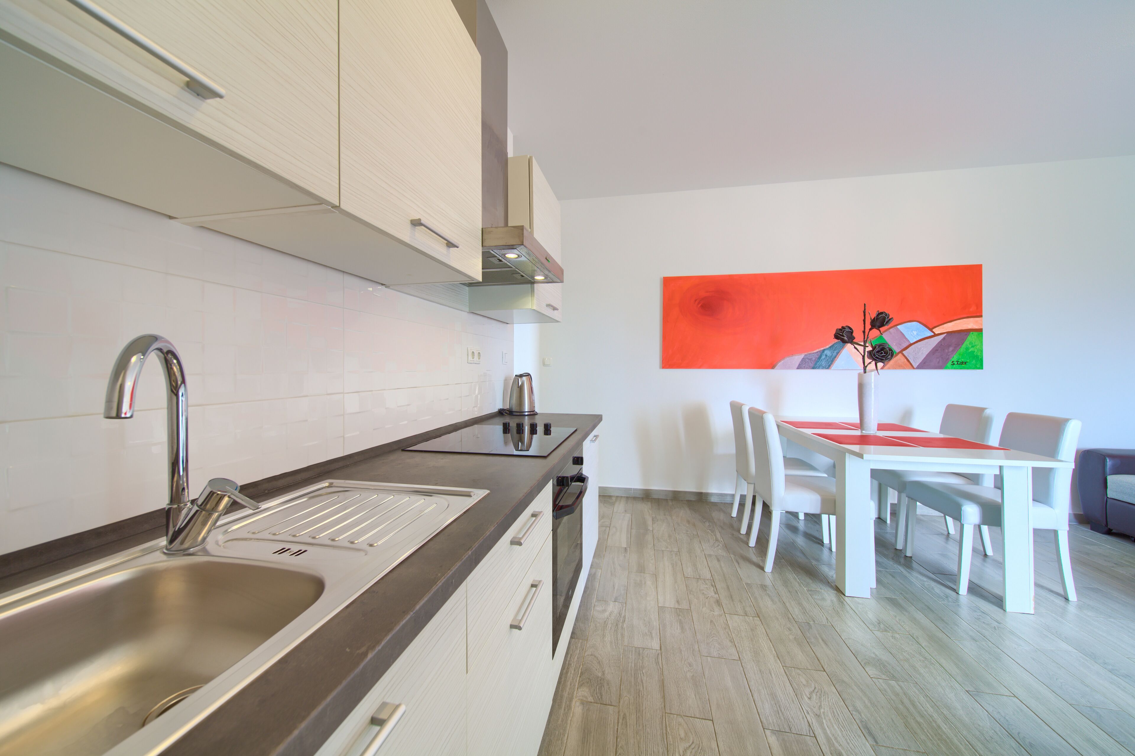 A modern kitchen with stainless steel appliances, a dining area with a white table and chairs, and a vibrant abstract painting on the wall.