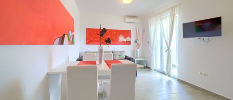 A modern living room with a dining area, featuring a large red artwork, a flat-screen TV, and a large window with curtains.