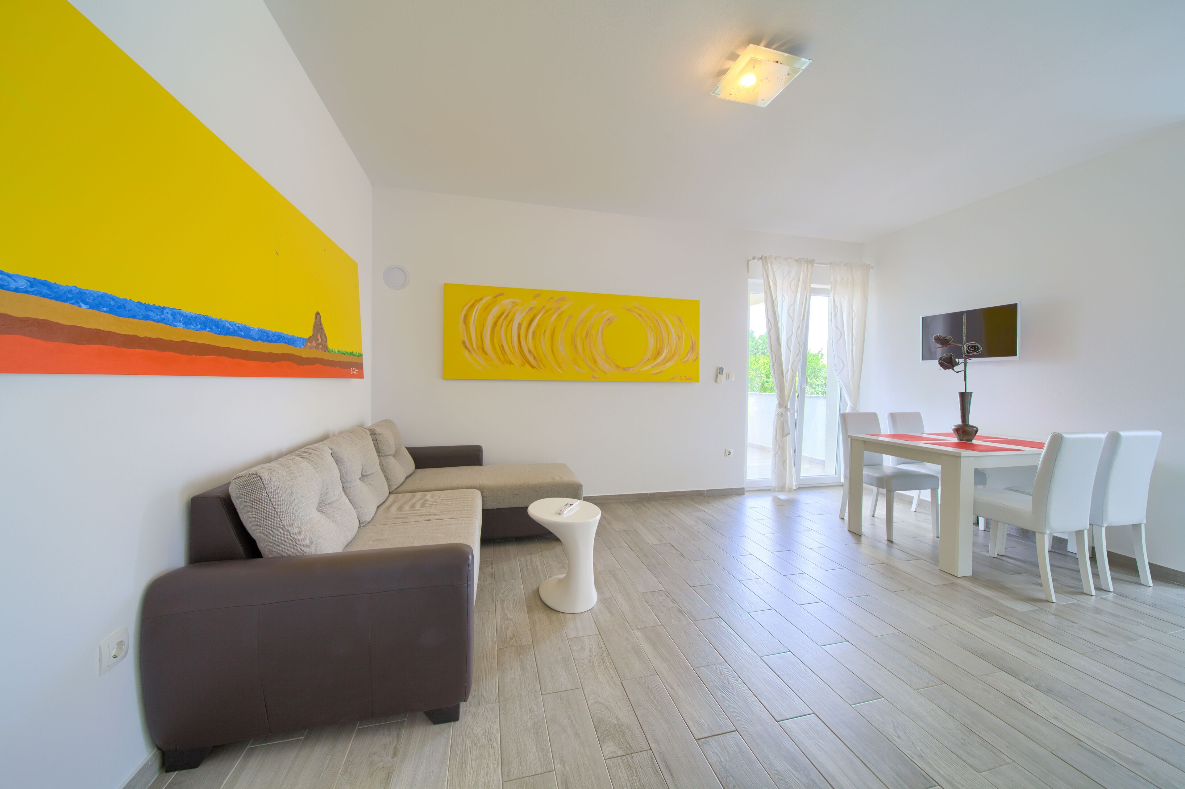 A modern living room with a sectional sofa, a dining table with chairs, and a TV on the wall.