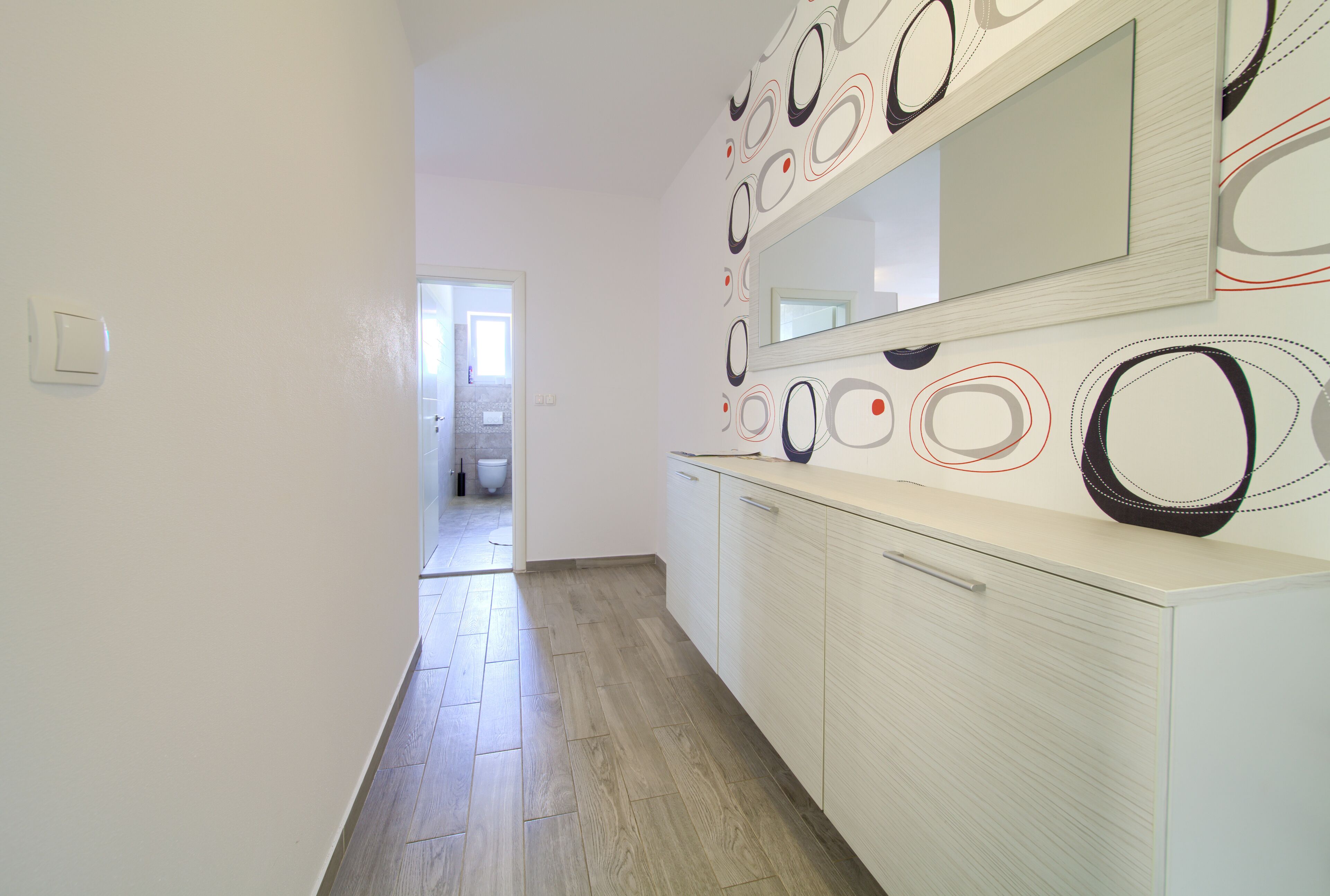 A hallway with a light-colored wooden floor, white walls, and a large mirror with abstract designs.