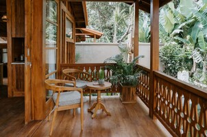 Un balcón de madera con mesa y sillas, rodeado de exuberante vegetación y una cerca decorativa.