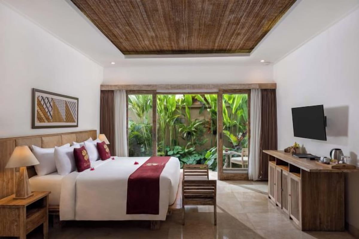 A hotel room with a bed, a TV, a desk, and a view of greenery outside.