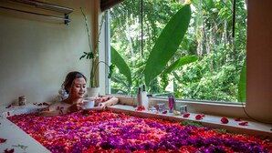 A bathtub filled with colorful flower petals, a window with a view of lush greenery, and a potted plant on the windowsill. - The Kalyana Ubud Resort (Ubud)