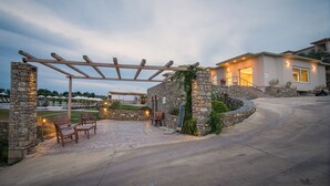 Een stenen pergola met houten stoelen en een bank, een stenen muur begroeid met groen, en een modern huis op de achtergrond.