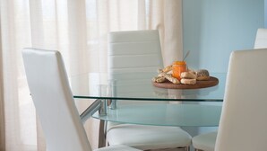 Een eettafel met glazen blad, witte stoelen, een glas water en een schaal met gebak en een pot jam.