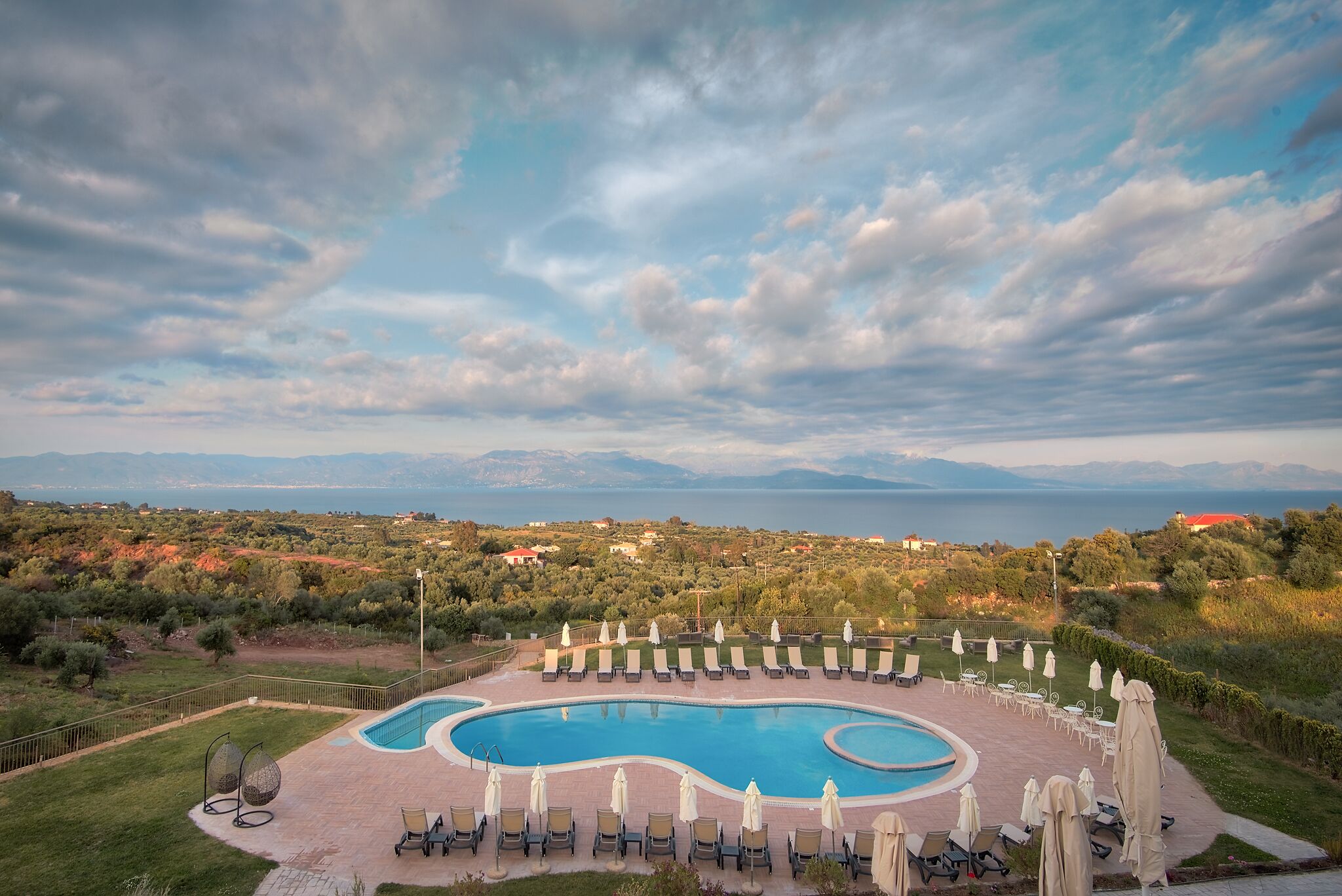 Ein Poolbereich mit Liegestühlen und Sonnenschirmen, umzäumt von einem Zaun, mit Blick auf das Meer und die Berge im Hintergrund.