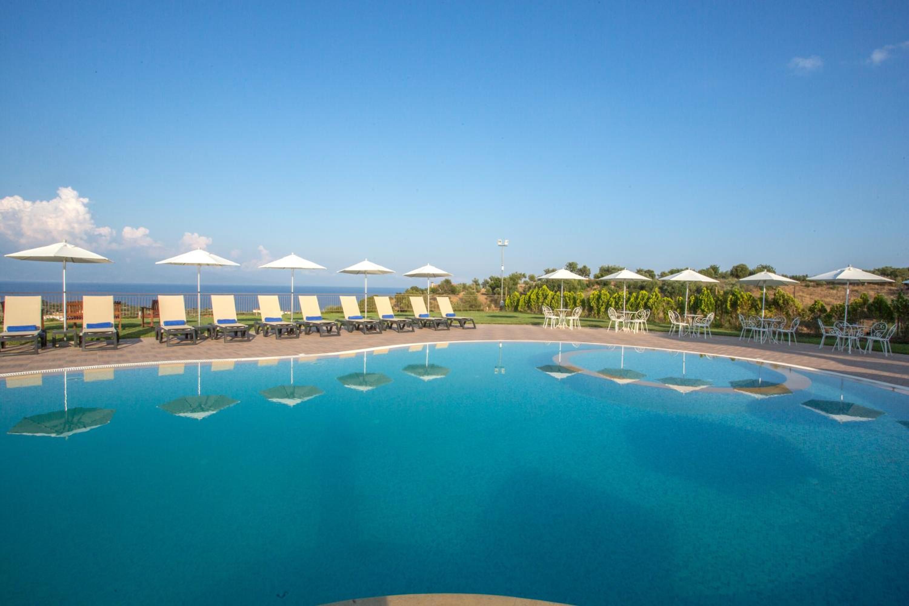 A pool with lounge chairs and umbrellas, surrounded by greenery and a clear sky.