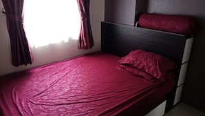 Un lit simple avec une couvre-lit rouge et un coussin assorti, une tête de lit et une fenêtre avec des rideaux.