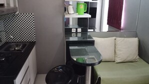 Une cuisine de taille réduite avec un plan de travail noir, un évier et une cuisinière. On aperçoit une tasse verte sur une étagère, et une fenêtre avec un rideau rouge.