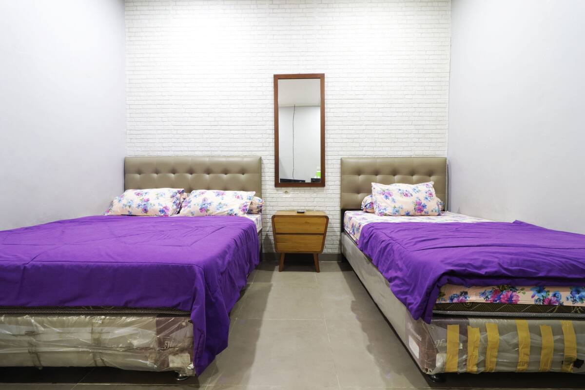 A room with two beds, each with a purple bedspread and floral pillows, a wooden nightstand, and a mirror on the wall.