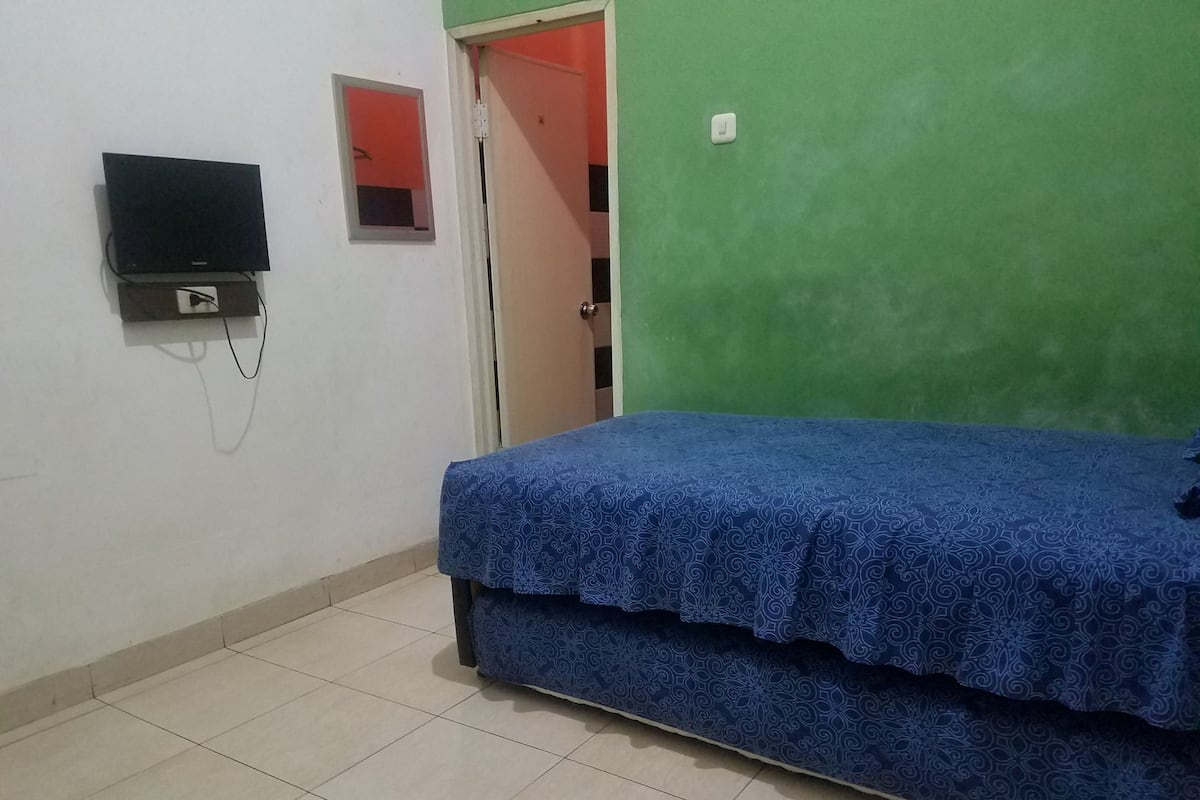 Una stanza con un letto, una televisione fissata al muro e una porta che conduce a un'altra stanza.
