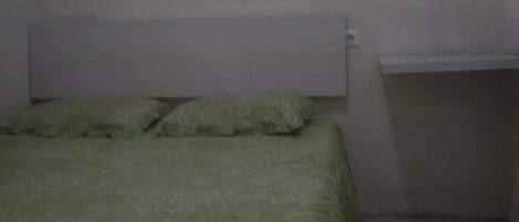 Una habitación pequeña y tenuemente iluminada, con una cama individual, un cabecero blanco y un pequeño estante adosado a la pared.