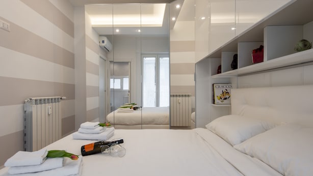 Ein modernes Hotelzimmer mit einem großen Bett, einem Nachttisch mit Flasche und Gläsern, einem Heizkörper und einem Spiegel, der den Raum reflektiert.