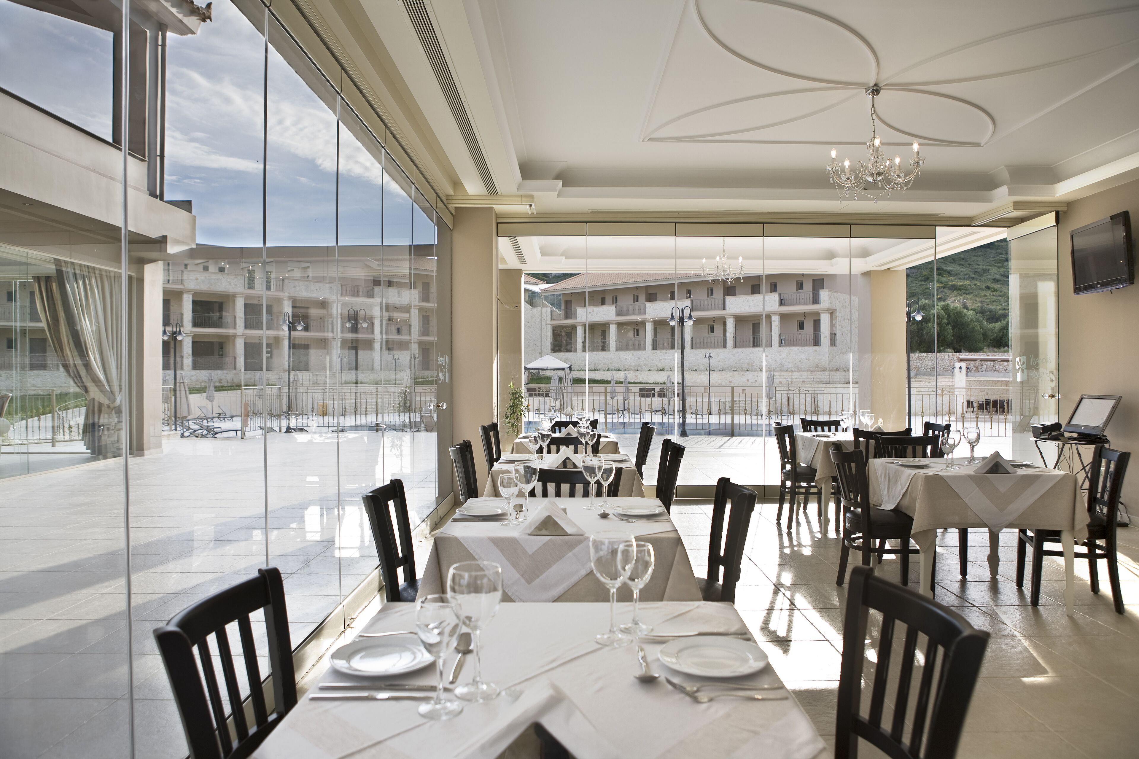 Un comedor con mesas dispuestas para una comida, amplias ventanas que ofrecen vistas a un edificio y un paisaje montañoso.