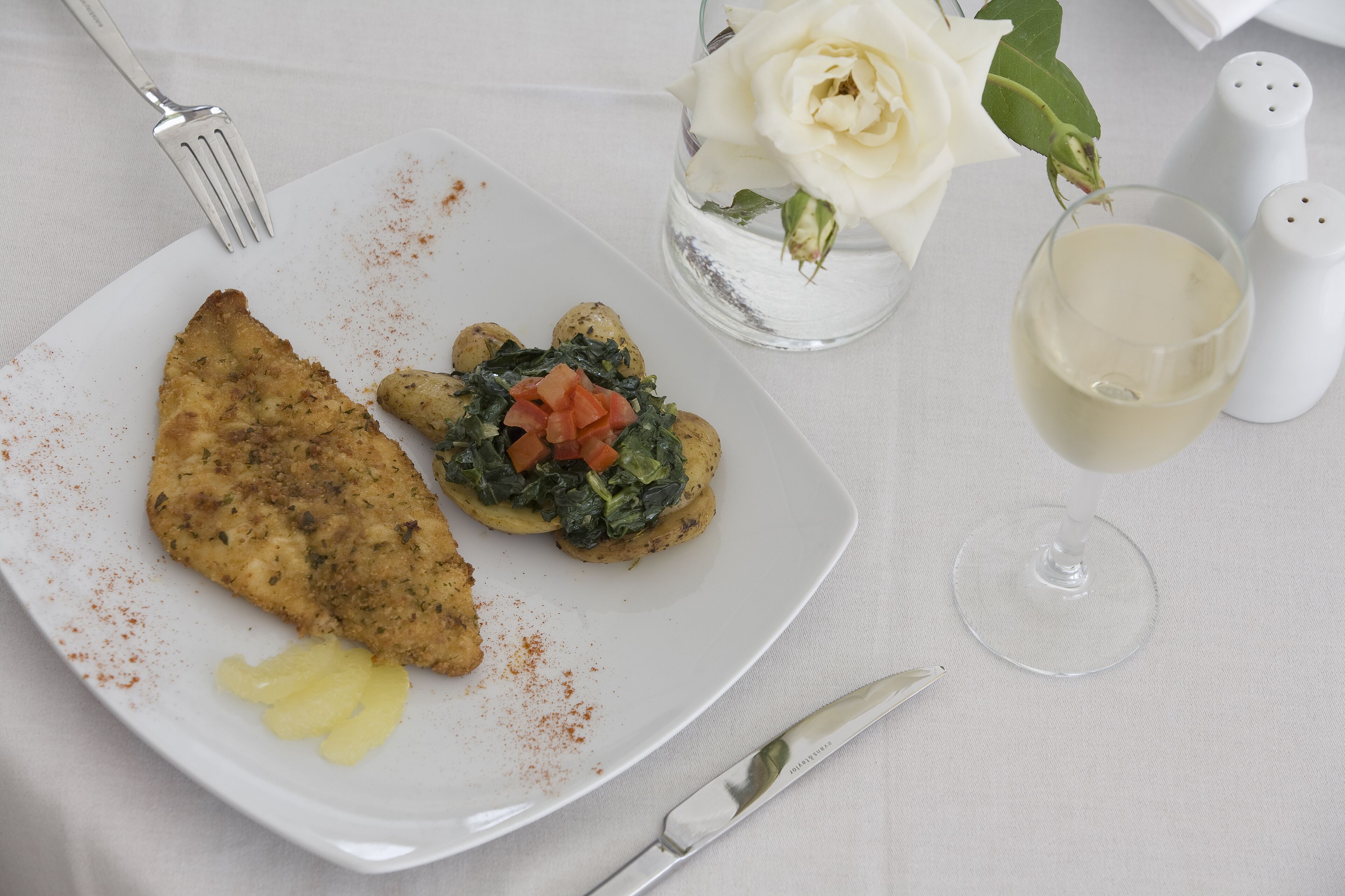 A plate with a piece of fried fish, a vegetable patty, and a slice of pineapple on a white plate. There is a glass of white wine and a white rose in a glass vase.