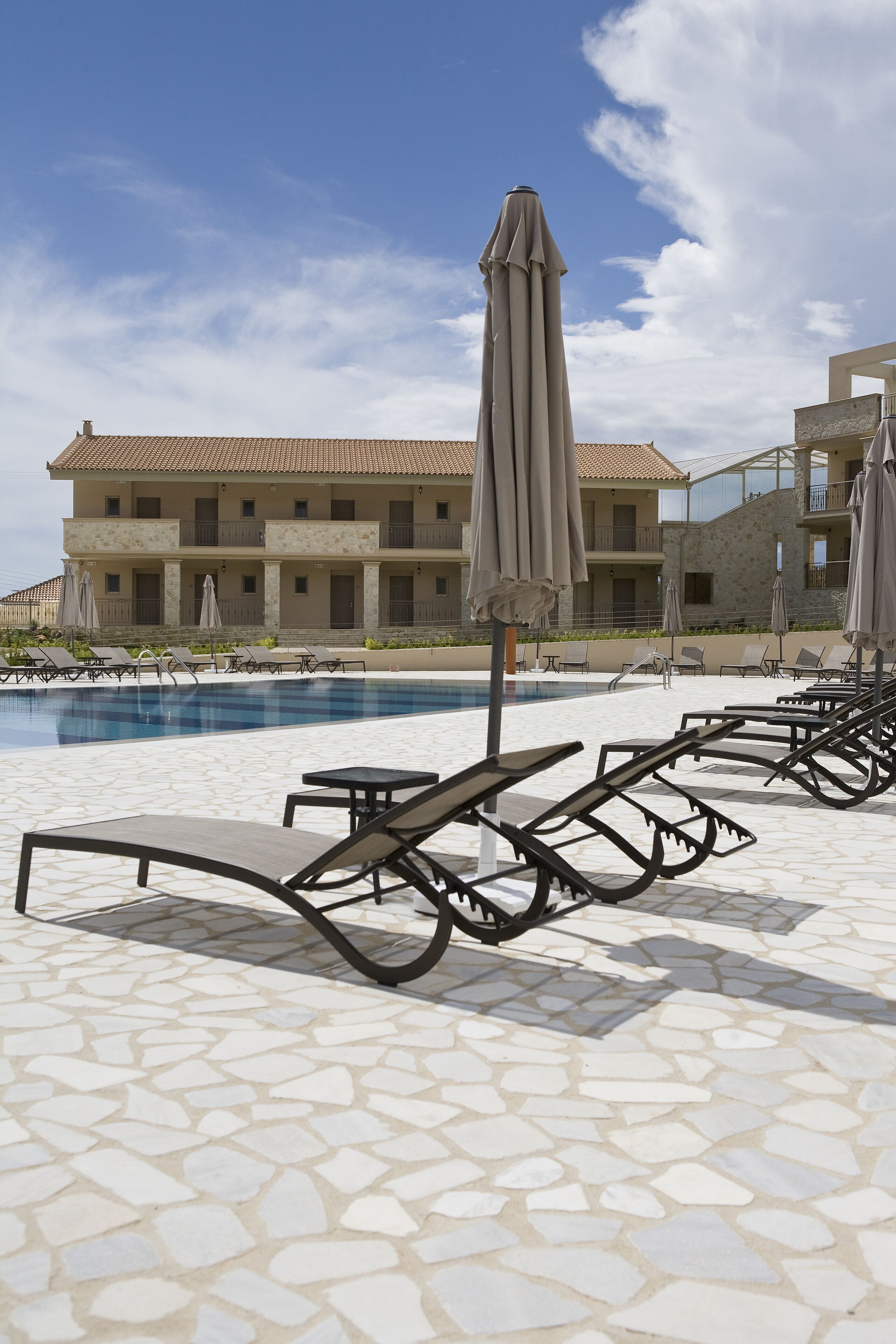 A poolside area with lounge chairs and umbrellas, a tiled floor, and a building with a tiled roof in the background.