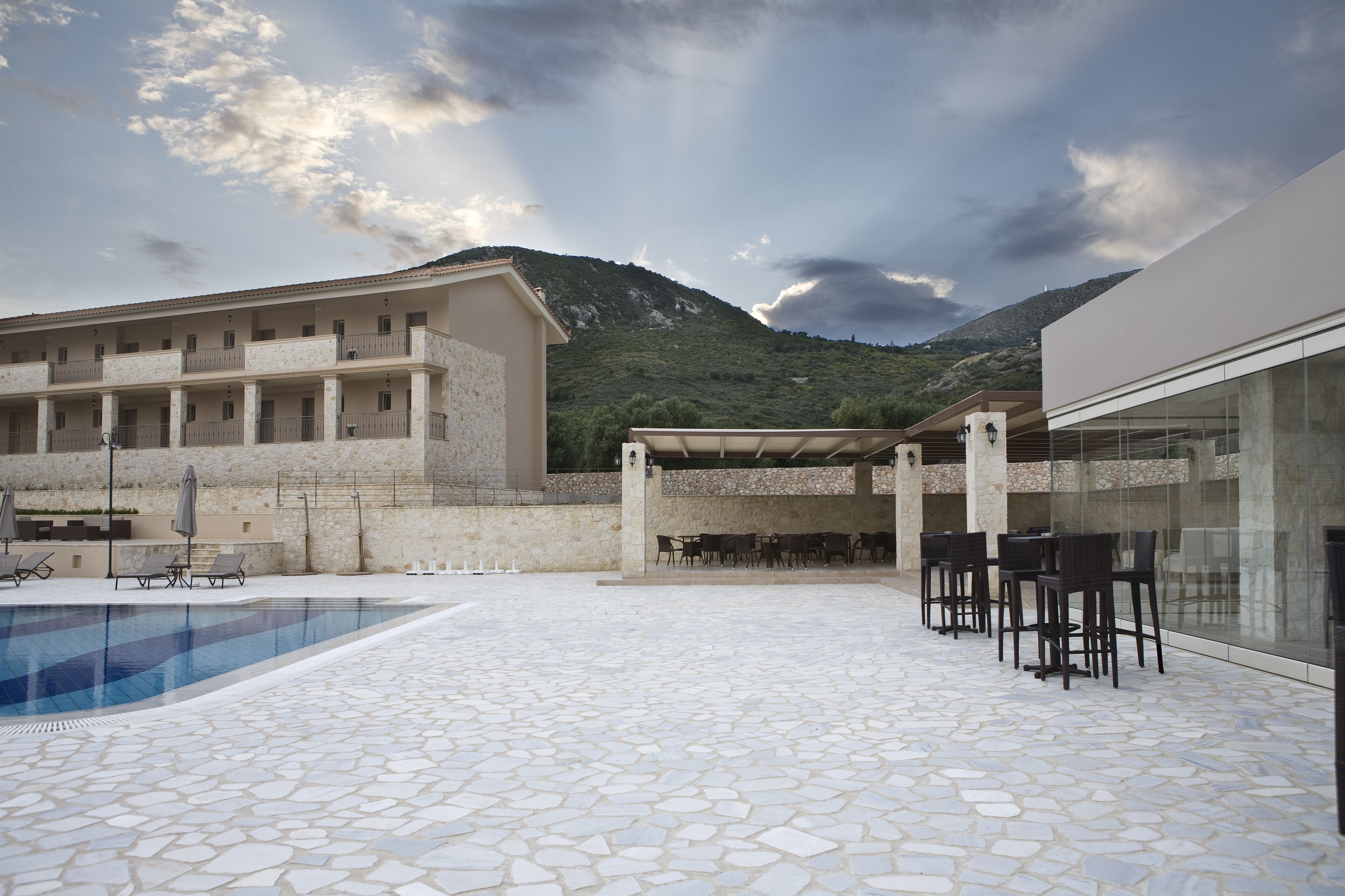 Zona de piscina con suelo de baldosas de piedra, mobiliario exterior y montañas al fondo.