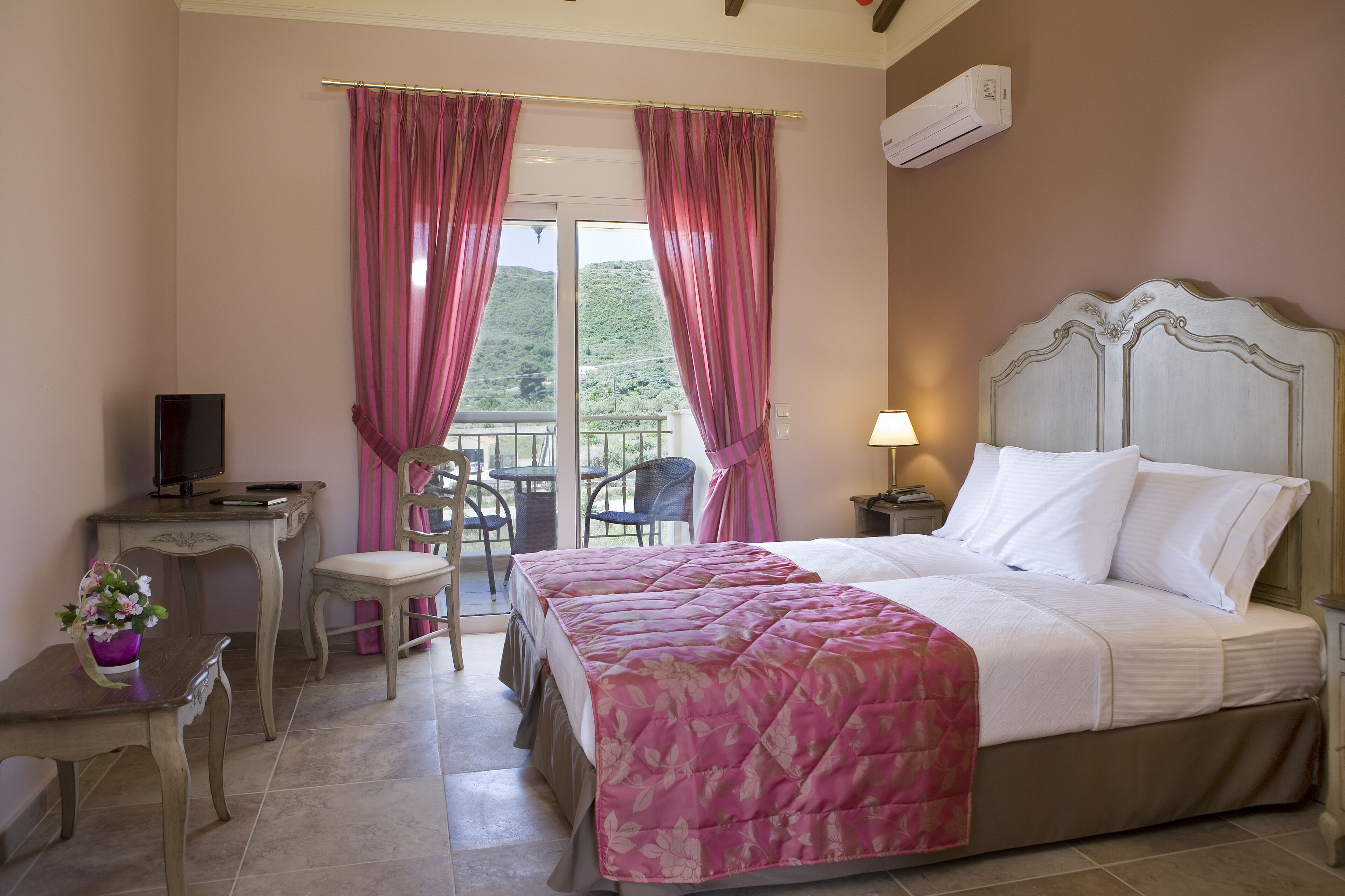 Une chambre à coucher avec un lit, un bureau avec une télévision, une chaise, une table avec un vase de fleurs et un balcon avec des rideaux.