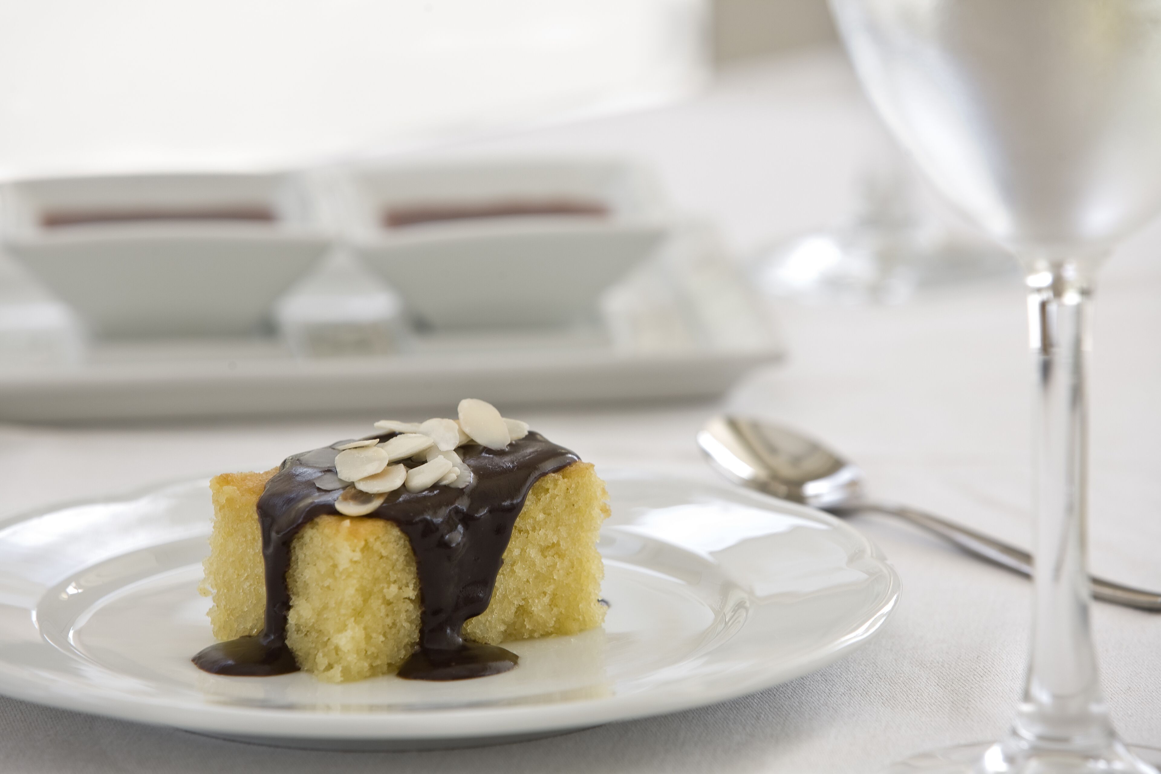 Une part de gâteau nappée de sauce au chocolat et parsemée d’amandes, présentée sur une assiette blanche.