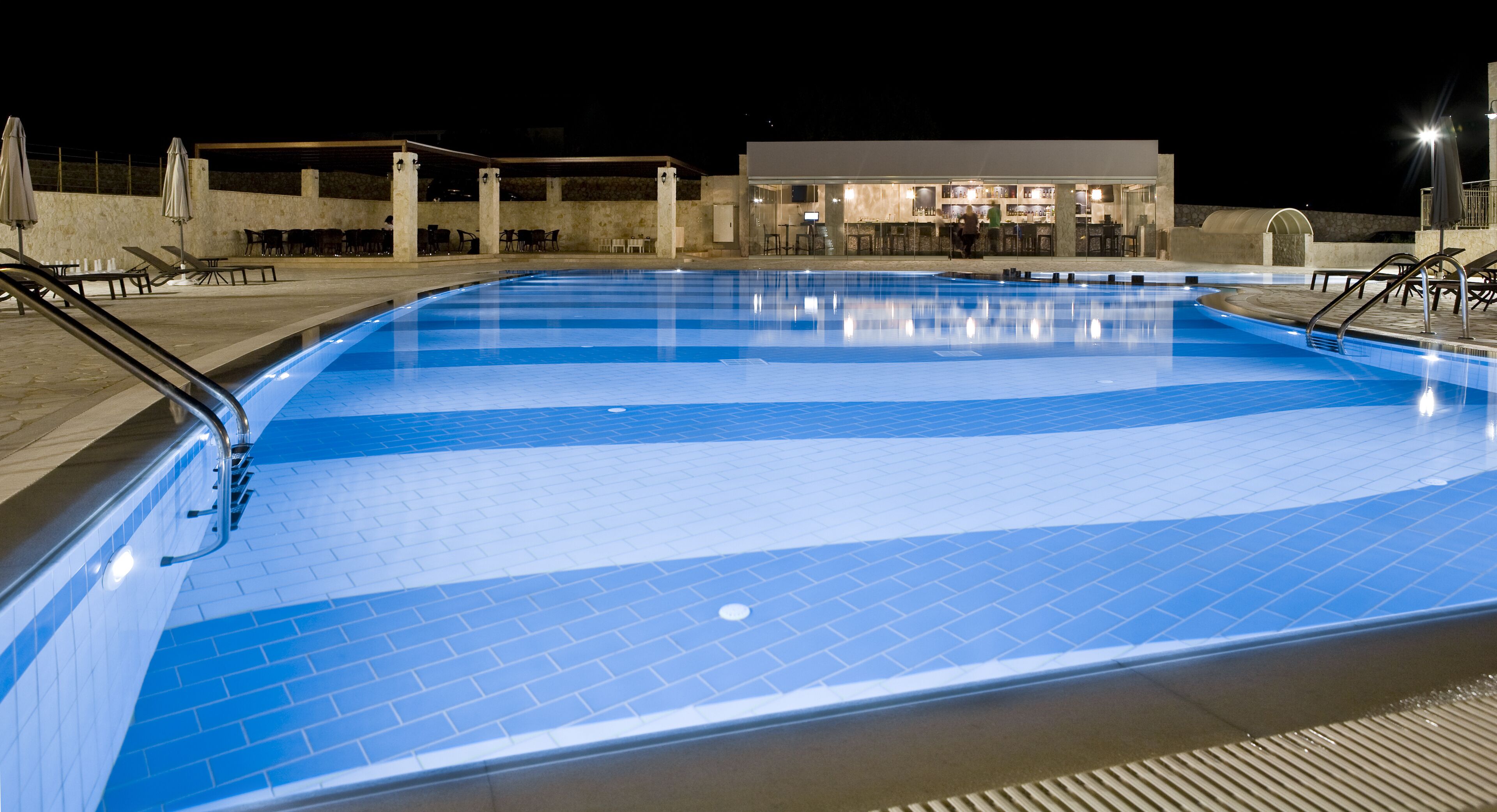 Piscina com deck ladrilhado, espreguiçadeiras e área de bar ao fundo.