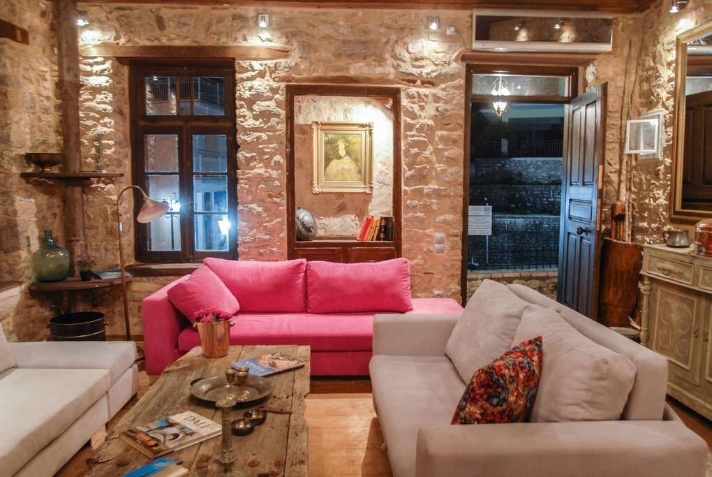 A cozy living room with a stone wall, a pink sofa, a white sofa, and a wooden coffee table with books and a lamp.